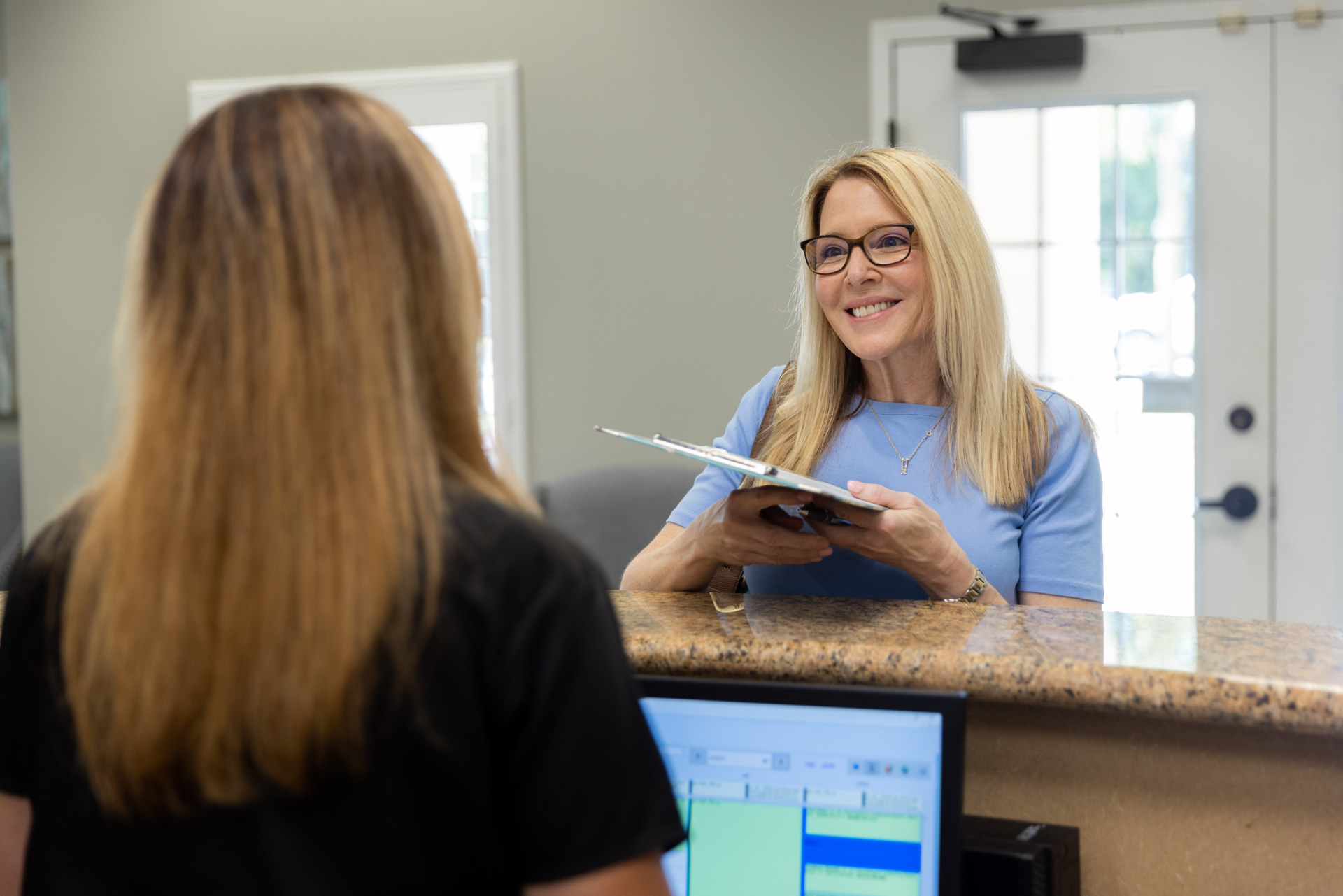 A lovely, mature blonde woman hands her introductory paperwork to a provider in our comfortable lobby before having her consultation. She has arrived to talk about having a non-surgical neck lift in Southlake, TX.